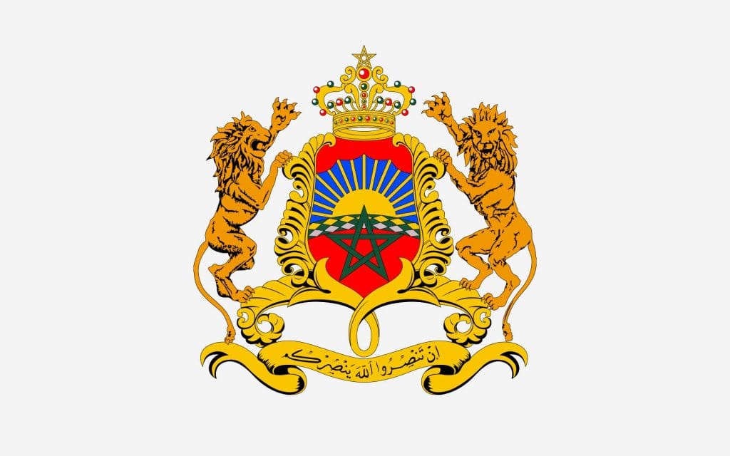 Emblème - Armoiries du Royaume du Maroc représentant deux lions dressés tenant un blason central. Le blason affiche un soleil levant, une étoile verte à cinq branches et un motif géométrique sur fond rouge. Il est surmonté d'une couronne royale richement ornée, avec une devise en arabe inscrite sur un ruban doré. L’ensemble symbolise la souveraineté, l’unité nationale et les valeurs royales du pays.
